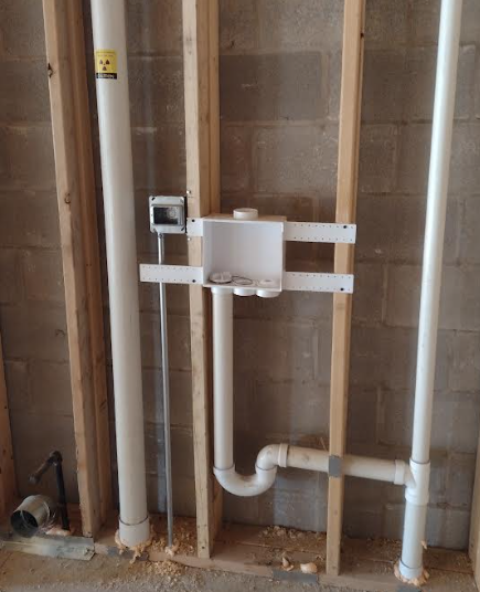 Field photo of laundry room “grey box” washer connection showing supply valves, waste line, trap, and PVC vent stack installed in framing.