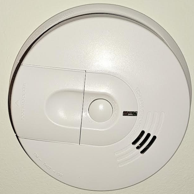 Close-up of a ceiling-mounted 120V interconnected smoke alarm, showing the battery backup compartment and test button.