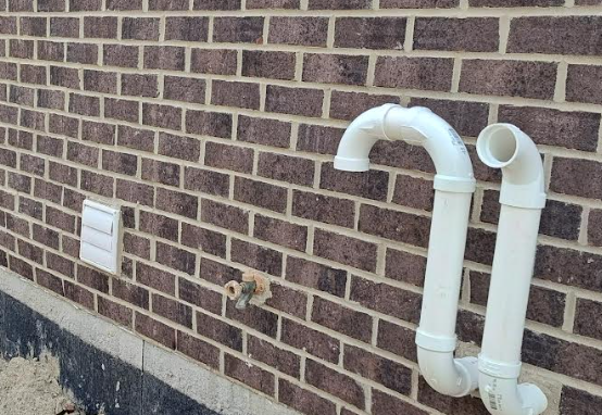 Code-compliant exterior dryer vent hood properly installed on a brick wall, with minimum clearance from grade and adjacent mechanical exhaust terminations.